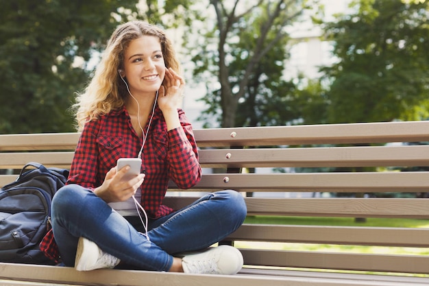 Hermosa mujer sonriente sentada y escuchando música en el teléfono inteligente en el banco al aire libre. Tecnología, redes sociales, educación y concepto independiente, espacio de copia