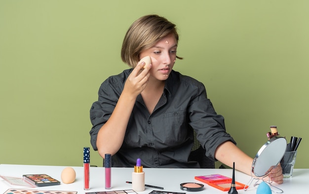 Hermosa mujer sonriente se sienta a la mesa con herramientas de maquillaje aplicando crema de tono con esponja mirando en el espejo