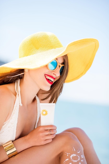 Hermosa mujer sonriente con sombrero amarillo tomando el sol sosteniendo una botella con protector solar loción protectora UV en la playa