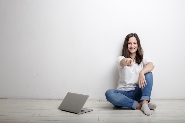 Hermosa mujer sonriente te señala con el dedo feliz sentado junto a la pared en el piso al lado del cuaderno abierto