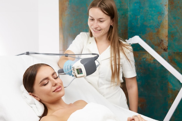 Hermosa mujer con sonrisa en la cara recibiendo tratamiento facial con láser en la clínica de belleza