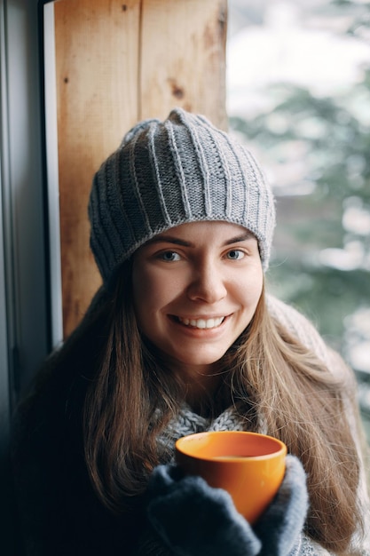 Hermosa mujer sosteniendo y bebiendo una taza de café o cacao con guantes sentada en casa junto a la ventana