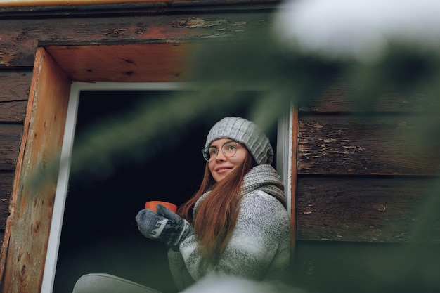 Hermosa mujer sosteniendo y bebiendo una taza de café o cacao con guantes sentada en casa junto a la ventana