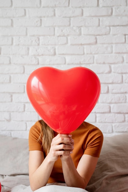 Hermosa mujer sosteniendo un globo con forma de corazón rojo sentada en la cama