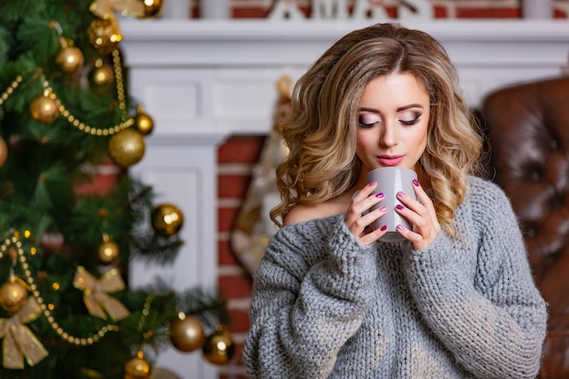 Hermosa mujer sosteniendo una taza de café caliente en sus manos al fondo de una chimenea blanca en la habitación con decoraciones de Año Nuevo y un árbol de Navidad