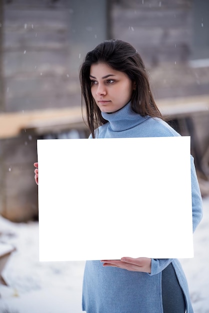 Hermosa mujer sostiene una tarjeta blanca en blanco Chica morena mientras nieve al aire libre Maqueta de arte