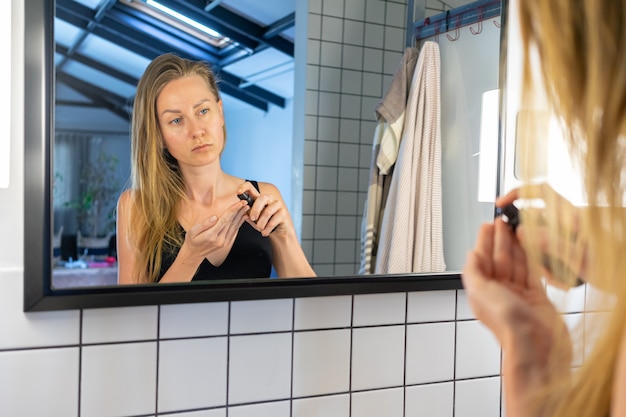 Hermosa mujer sostiene un tarro de crema aplicando crema hidratante para la piel de la cara, de pie frente al espejo del baño.