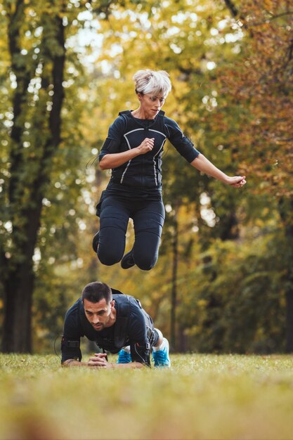 Una hermosa mujer y su alegre pareja están haciendo ejercicios en el parque, vestidos con un traje negro con un simulador electrónico EMS para estimular sus músculos.