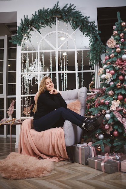 hermosa mujer con un suéter de punto se sienta en un sillón con el telón de fondo de una habitación lujosamente decorada con un árbol de Navidad. Buen espíritu de año nuevo