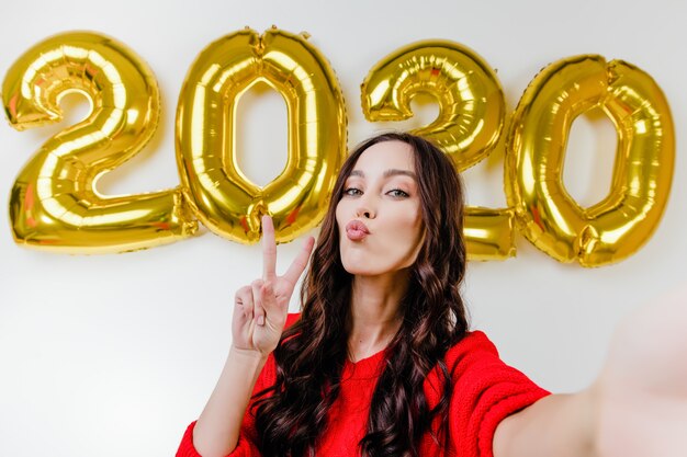 Hermosa mujer en suéter rojo haciendo selfie divertido frente a 2020 globos de año nuevo