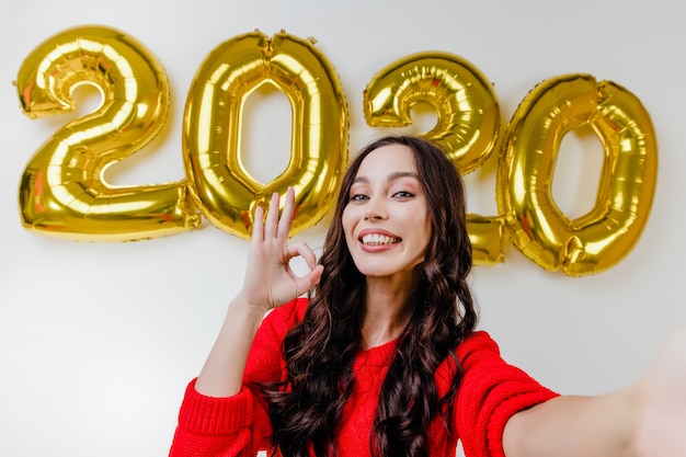 Hermosa mujer en suéter rojo haciendo selfie divertido frente a 2020 globos de año nuevo