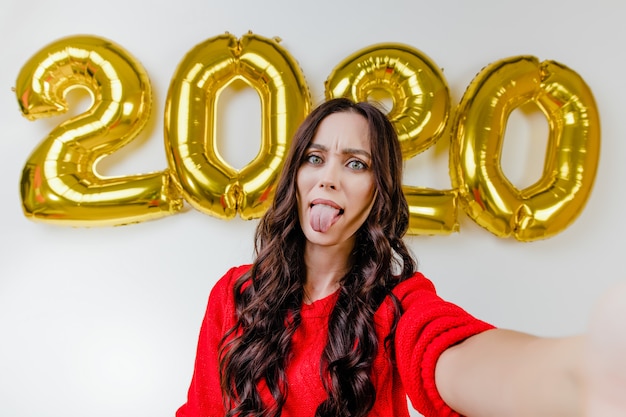 Hermosa mujer en suéter rojo haciendo selfie divertido frente a 2020 globos de año nuevo