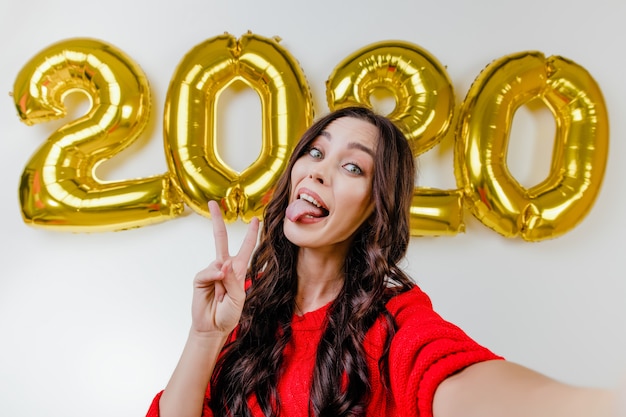 Hermosa mujer en suéter rojo haciendo selfie divertido frente a 2020 globos de año nuevo