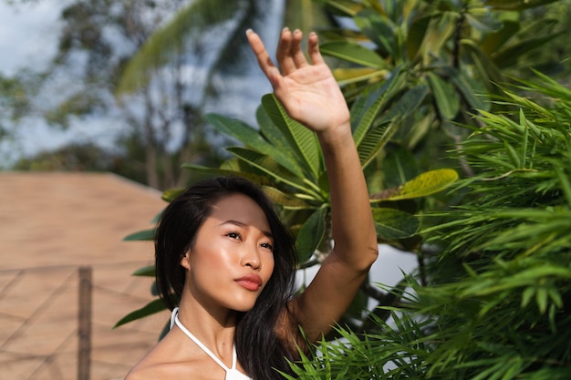 Hermosa mujer tailandesa asiática con piel bronceada oscura suave y limpia posando al aire libre en vestido blanco con fondo tropical