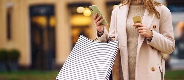 Foto hermosa mujer con teléfono y una tarjeta de crédito chica rubia en un abrigo ligero con bolsas de compras