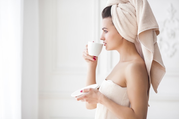 Hermosa mujer en la toalla después de bañarse tomando café por la mañana