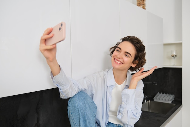 Hermosa mujer se toma selfie en un teléfono inteligente posando en la cocina y sonriendo a la cámara del móvil para tomar una foto