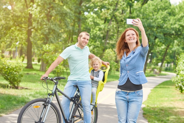 Hermosa mujer tomando selfie con esposo y bebé en bicicleta mientras se relaja al aire libre en el parque.