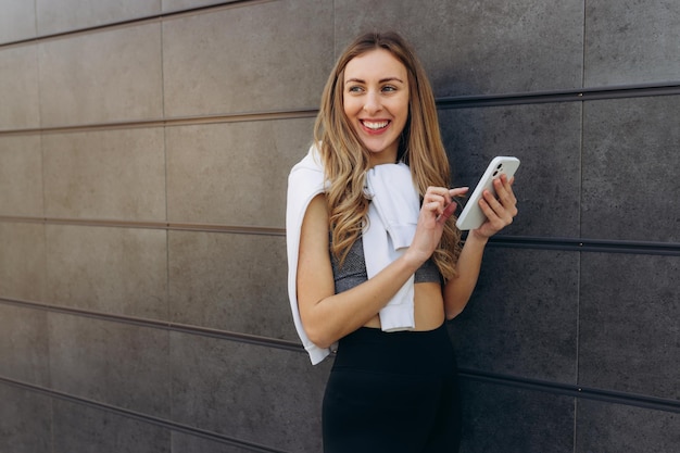 Hermosa mujer con top deportivo gris mientras sostiene el teléfono inteligente en las manos con cabello claro usando el teléfono celular mientras posa al aire libre cerca de la pared gris Copiar espacio