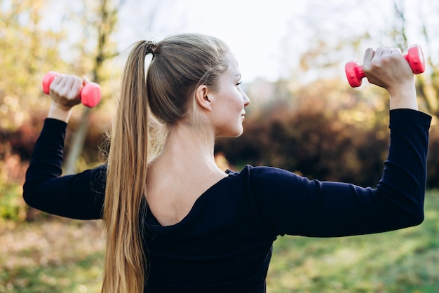 Hermosa mujer trabajando al aire libre. Deportiva mujer entrenando con pesas, vista posterior