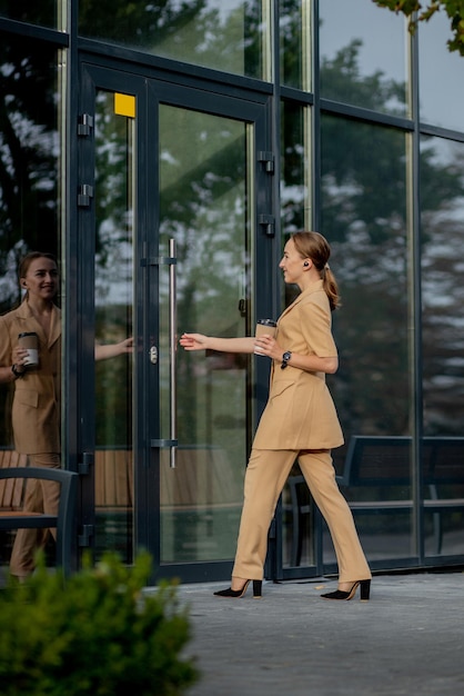 Hermosa Mujer Va A Trabajar Con Café Caminando Cerca Del Edificio De Oficinas. Retrato de mujer de negocios exitosa sosteniendo una taza de bebida caliente en la mano en su camino al trabajo en la calle de la ciudad. Alta resolución.