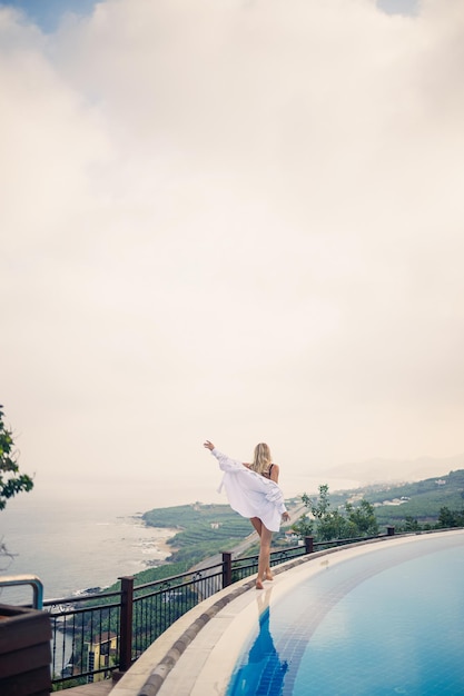 Hermosa mujer en traje de baño negro posando cerca de la piscina al aire libre con vista al mar