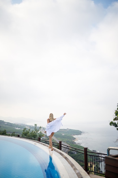 Hermosa mujer en traje de baño negro posando junto a la piscina al aire libre con vistas al mar