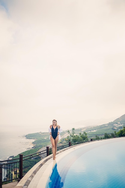 Hermosa mujer en traje de baño negro posando junto a la piscina al aire libre con vistas al mar