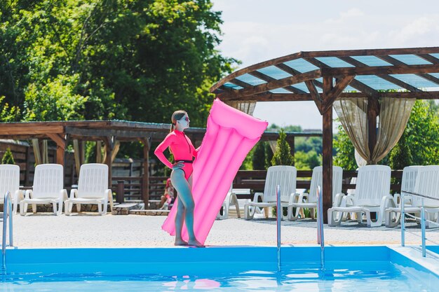 Una hermosa mujer en traje de baño rosa y con un colchón inflable se encuentra cerca de una gran piscina Vacaciones de verano en un hotel con piscina Chica joven en traje de baño y gafas de sol