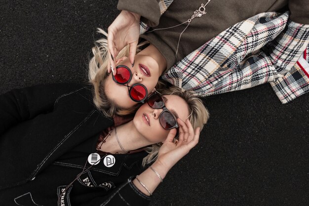 Hermosa mujer urbana con cabello rubio en ropa negra con gafas de sol se encuentra cerca de la novia moderna en ropa vintage en gafas rojas