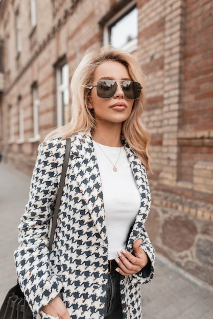 Hermosa mujer urbana con gafas de sol en chaqueta de moda con bolso camina por la calle Thea cerca de un edificio de ladrillo