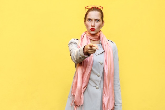 La hermosa mujer, vestida con un abrigo de otoño, abriendo la boca ampliamente, habiendo sorprendido miradas de asombro, señalando con el dedo a la cámara. Foto de estudio, pared amarilla