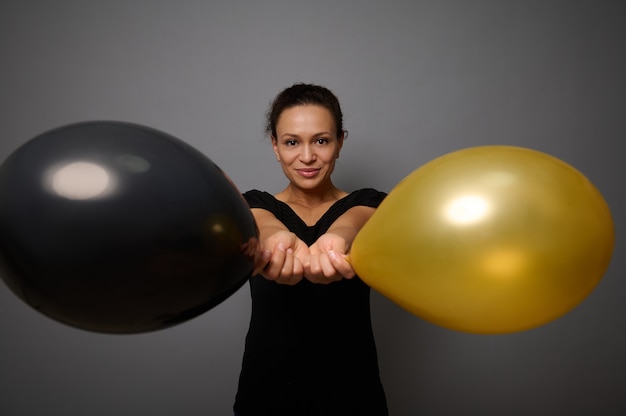 Hermosa mujer vestida con sonrisas negras mirando a la cámara y posa sobre un fondo gris sosteniendo globos de aire dorados y negros en su mano extendida. Concepto de viernes negro con espacio de copia para anuncios