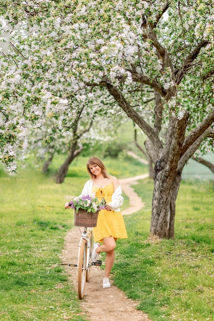 Foto una hermosa mujer con un vestido amarillo y una bicicleta camina por un jardín en flor en primavera.