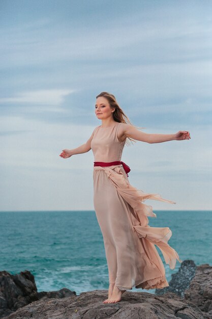 Hermosa mujer con un vestido beige se encuentra en una costa rocosa