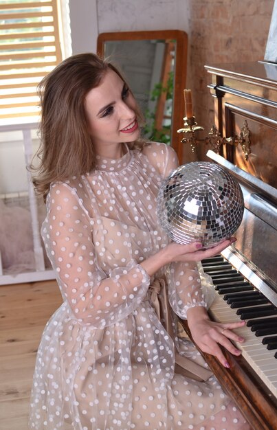 Hermosa mujer con un vestido beige sostiene una bola de discoteca en la mano y toca un piano retro