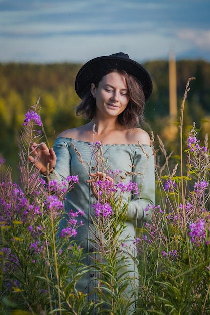 Foto hermosa mujer en vestido de belleza, sombrero negro con gran ramo de flores púrpuras se ríe y camina al aire libre en un prado durante el atardecer. concepto de estilo de vida.