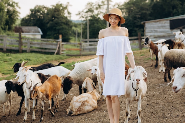 Hermosa mujer y vestido blanco y con un sombrero beige entre cabras en una granja ecológica