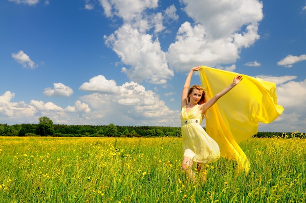 Hermosa mujer en un vestido de cóctel amarillo posando en un campo verde con seda de ligereza en sus manos