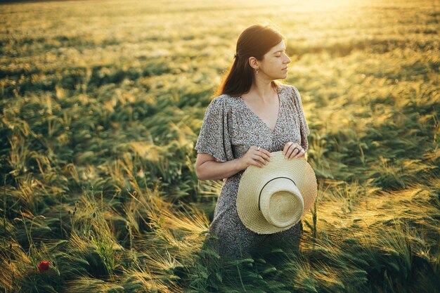 Hermosa mujer en vestido floral de pie en el campo de cebada a la luz del atardecer Momento tranquilo atmosférico Vida lenta rústica Mujer elegante sosteniendo sombrero de paja y disfrutando del campo de verano por la noche