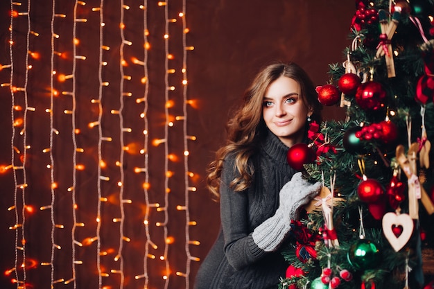 Hermosa mujer en vestido gris de pie contra el árbol de Navidad decorado.