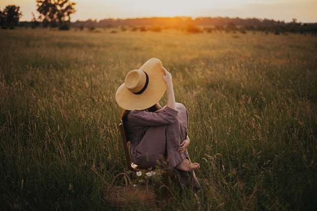 Hermosa mujer con vestido de lino y sombrero sentada en una silla en la pradera de verano disfrutando del atardecer Tranquilo