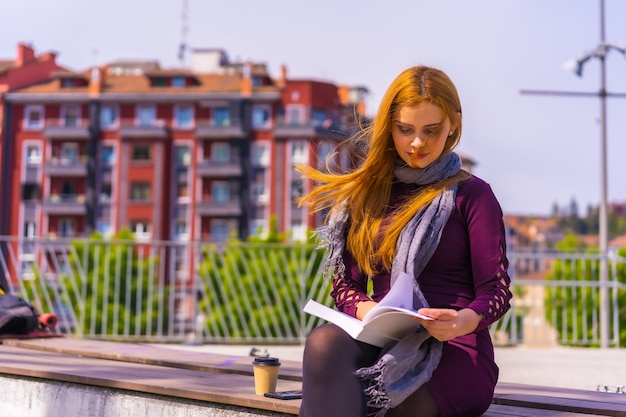 Hermosa mujer en vestido morado y bufanda leyendo un libro en un parque de la ciudad, imaginando y disfrutando de la lectura