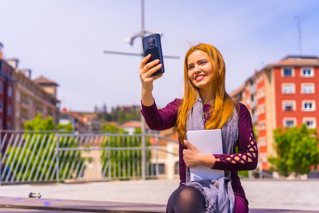 Hermosa mujer en vestido morado y bufanda con un libro en la mano en un parque de la ciudad, tomando un selfie con el teléfono móvil