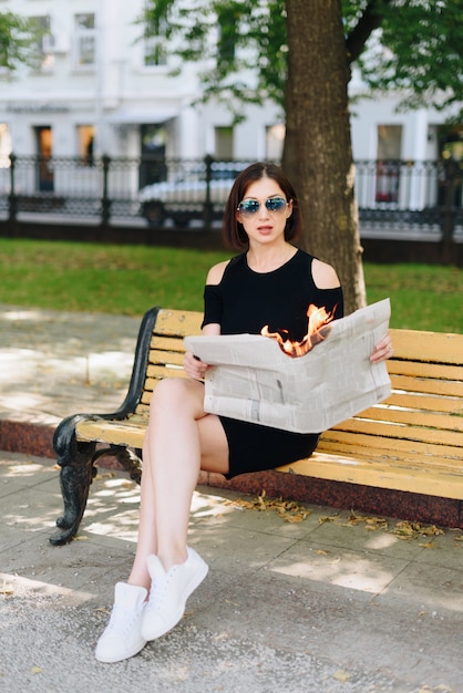 Hermosa mujer con un vestido negro en un parque con un periódico en llamas en sus manos