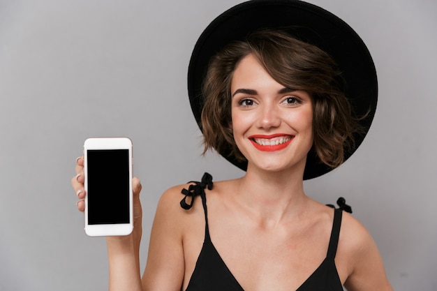 Hermosa mujer con vestido negro y sombrero sosteniendo smartphone, aislado sobre pared gris