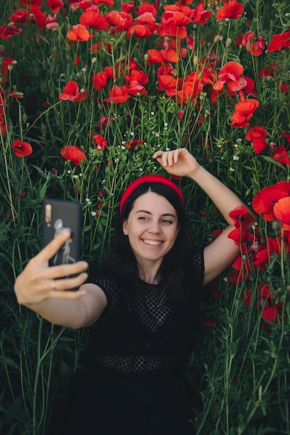 Hermosa mujer en vestido negro tomando selfie en el campo de flores de amapolas