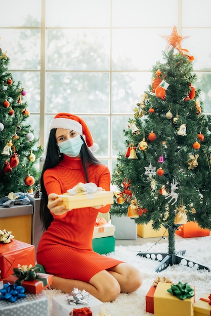 Una hermosa mujer con un vestido rojo y un gorro de Papá Noel con una mascarilla quirúrgica está sentada rodeada de coloridas cajas de regalo y disfruta de la Navidad sola en casa.