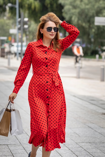 Hermosa mujer en vestido rojo de verano caminando alegremente sonriendo por la calle mientras hace compras llevando sus bolsas.