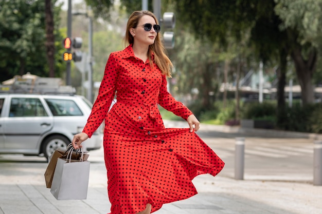 Hermosa mujer en vestido rojo de verano caminando alegremente sonriendo por la calle mientras hace compras llevando sus bolsas.